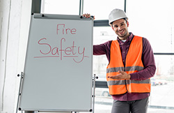 New Jersey fire alarm installation technician wearing blaze orange safety vest and whit hard hat standing next to display showing the words "Fire Safety"